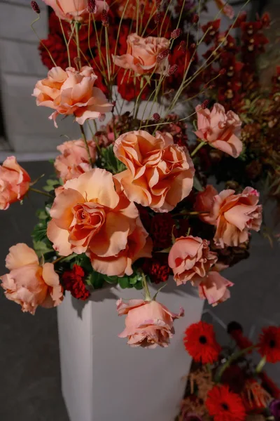 Wedding floral arrangement of peach rose centerpiece with burgundy blooms and greenery on a pedestal stand against a gray stone wall