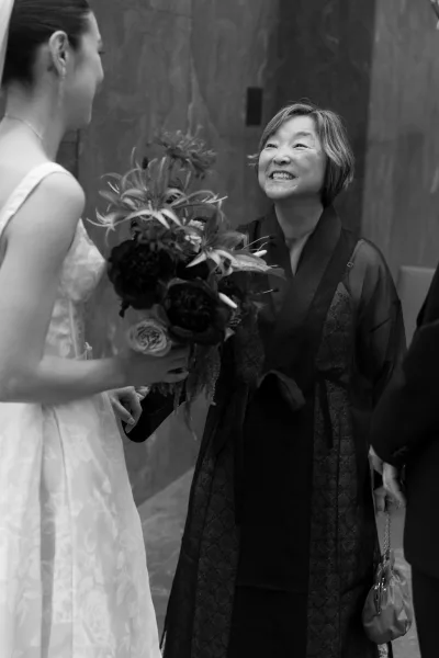 Wedding guest moment as the bride greets a guest while holding a textured bouquet, earrings and handbag visible against a wood panel wall indoors