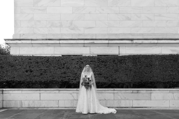 Bridal portrait in black and white of a bride holding bouquet, wearing a cathedral veil and necklace against a marble wall with hedges