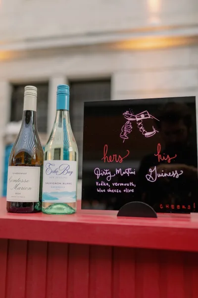 Wedding bar sign with signature drinks menu on a chalkboard, flanked by wine bottles on the bar counter outside by windows
