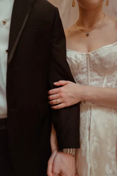 Couple portrait of bride holding groom’s arm, showing strapless wedding dress and black tuxedo, with veil against neutral wall