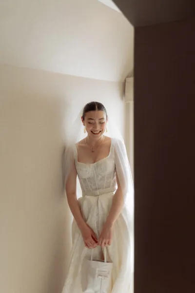 Bridal portrait of a smiling bride in a strapless wedding dress and veil, holding a mini purse by window light with soft shadows