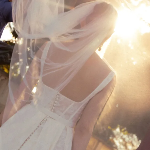 Bridal dress back with corset back wedding dress buttons and lace trim, bridal veil draped over as sun flare filters through trees outdoors