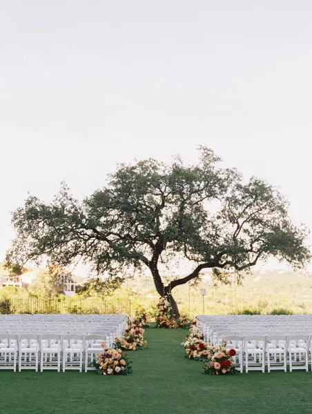 Ceremony setup with outdoor wedding ceremony seating of white folding chairs, aisle florals and greenery under a large tree with string lights