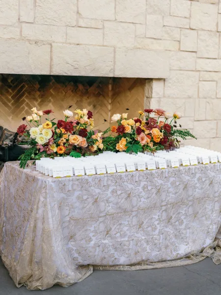 Escort card table with wedding escort cards set on a lace tablecloth, framed by a floral arrangement beside a stone fireplace backdrop