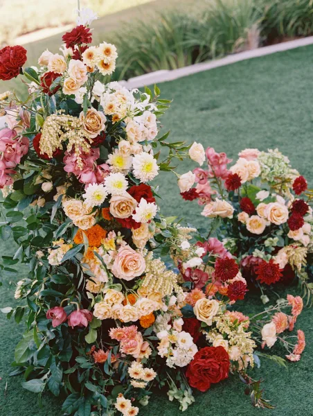 Wedding floral arrangement with roses and dahlias in an asymmetrical ground display beside a grass lawn pathway curb with greenery accents