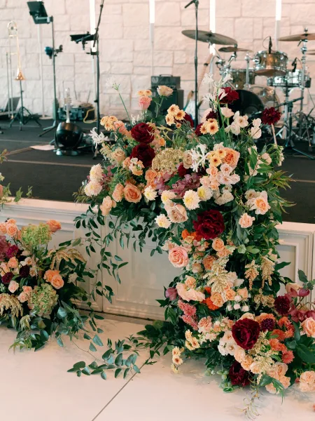 Wedding stage florals with ceremony stage flowers in lush rose and dahlia garlands on a riser beside drum set and mic stands, stone wall backdrop