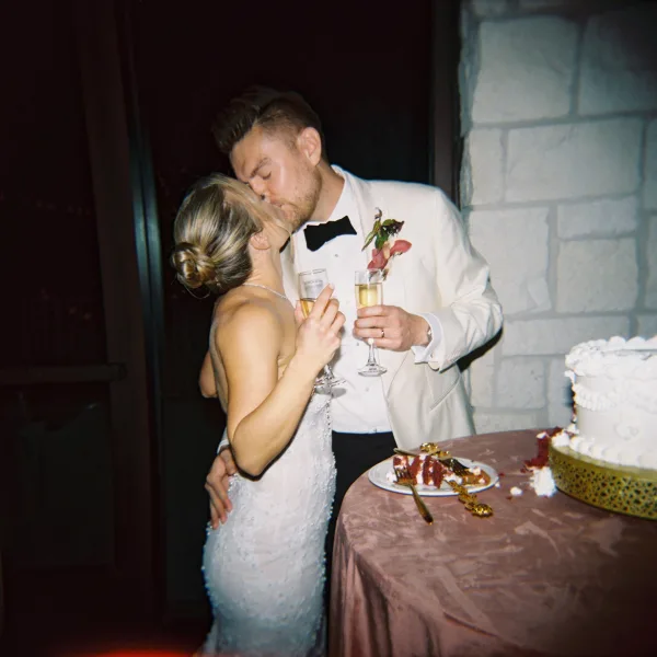 Wedding kiss beside a cake cutting moment table, couple holding champagne flutes in front of a stone wall and dark doorway