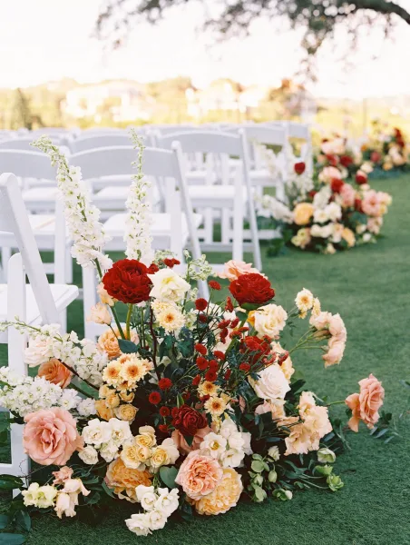 Ceremony aisle decor with outdoor wedding ceremony chairs lined in white, grounded by lush rose and ranunculus clusters on a grassy lawn