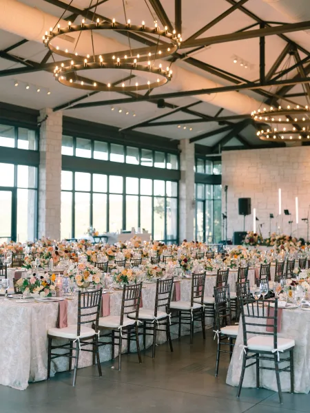 Reception tablescape with wedding reception table decor, pastel floral centerpieces and candles on ivory linens beneath round chandeliers in an industrial loft venue