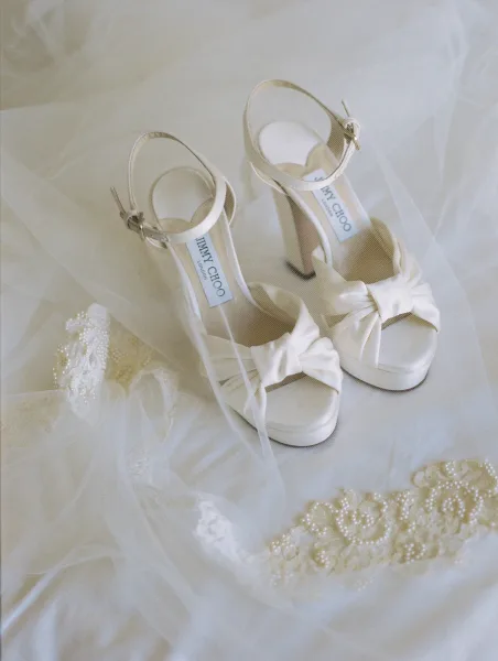 Bridal shoes, white wedding heels with bow detail and ankle straps, arranged on tulle veil with pearl-beaded lace on white fabric surface