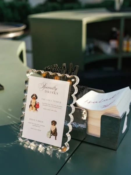 Bar menu sign with wedding signature drink menu in an acrylic holder on a bar cart with black straws and napkins, greenery behind