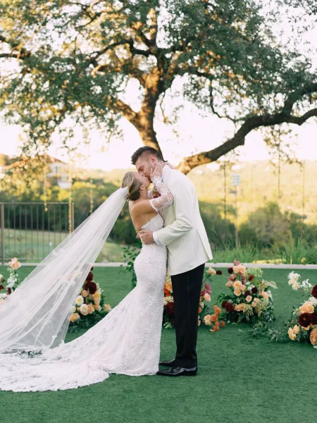 Wedding kiss as bride and groom kissing under a large tree, her long veil trailing over lace dress while he hugs her on the lawn