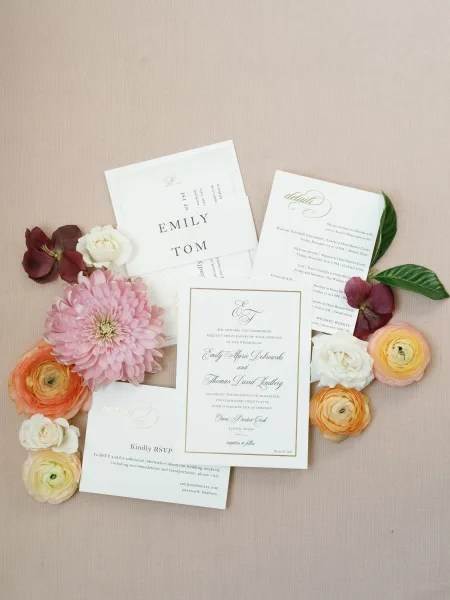 Invitation suite flat lay with wedding invitation cards, RSVP and details card, gold foil lettering, florals and greenery on neutral fabric