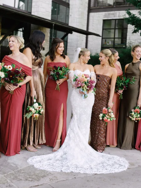 Bridesmaid group photo of bride with bridesmaids holding bouquets, bride’s cathedral veil and long train outside a stone building