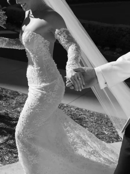 Couple portrait of bride and groom holding hands, her long veil and lace off-shoulder dress flowing along a garden stone path