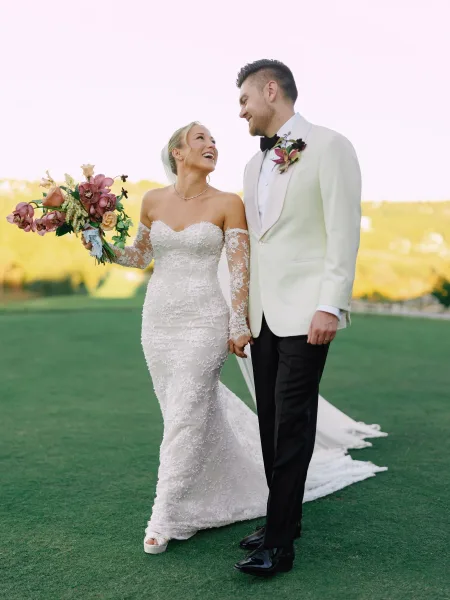 Couple portrait of bride and groom holding hands, her pink orchid bouquet and lace gown on a golf course lawn with hills beyond