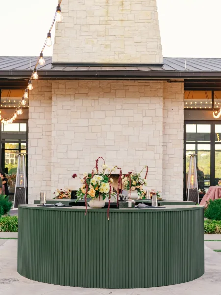 Wedding bar setup with floral arrangements and vases, lit by string lights beside a stone fireplace wall on an outdoor patio