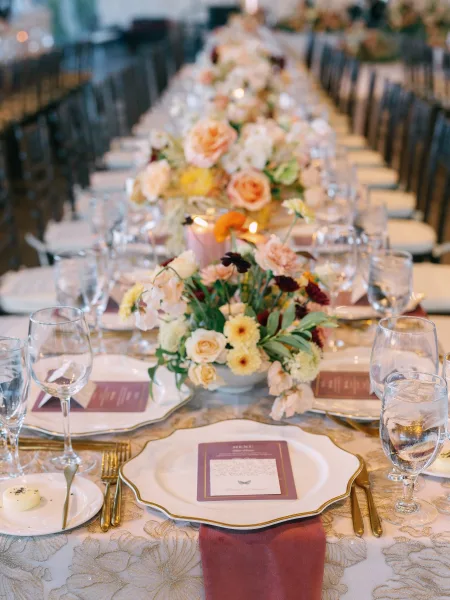Reception tablescape with taper candles, pastel floral runner, gold rim plates and glassware on a long banquet table in soft indoor light