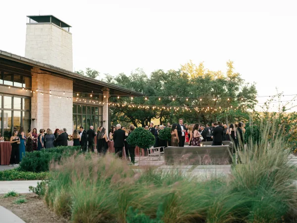 Wedding cocktail hour on a patio with string lights, cocktail tables and lounge seating outside a stone building with greenery and trees