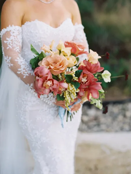 Bridal bouquet of coral and peach roses with orchid blooms and greenery, held against a white lace off-the-shoulder dress outdoors by a stone wall