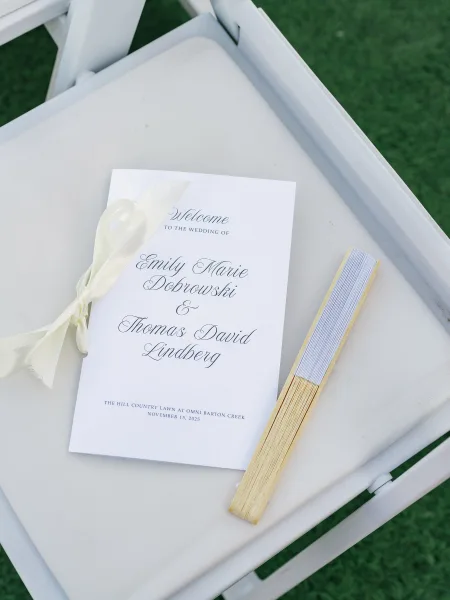 Wedding program with calligraphy print tied with an ivory ribbon beside a handheld fan on a white folding chair on green grass lawn