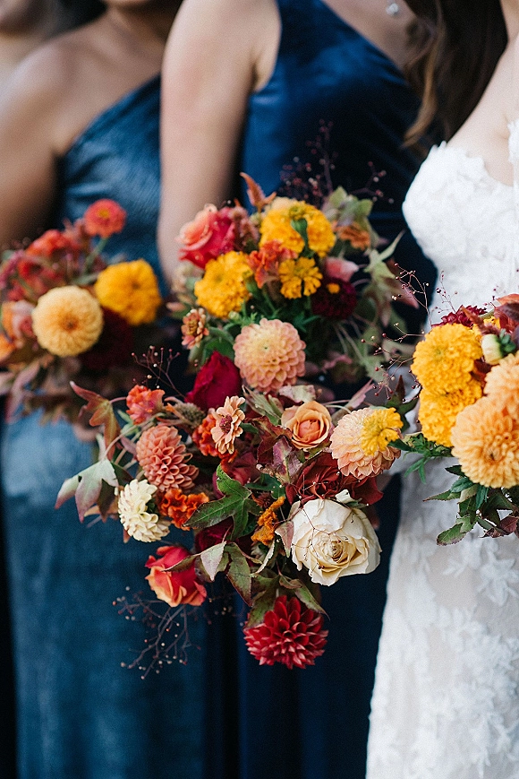 Bridesmaid bouquets of dahlias and roses in warm fall tones, held by bridesmaids in navy dresses beside the bride in lace