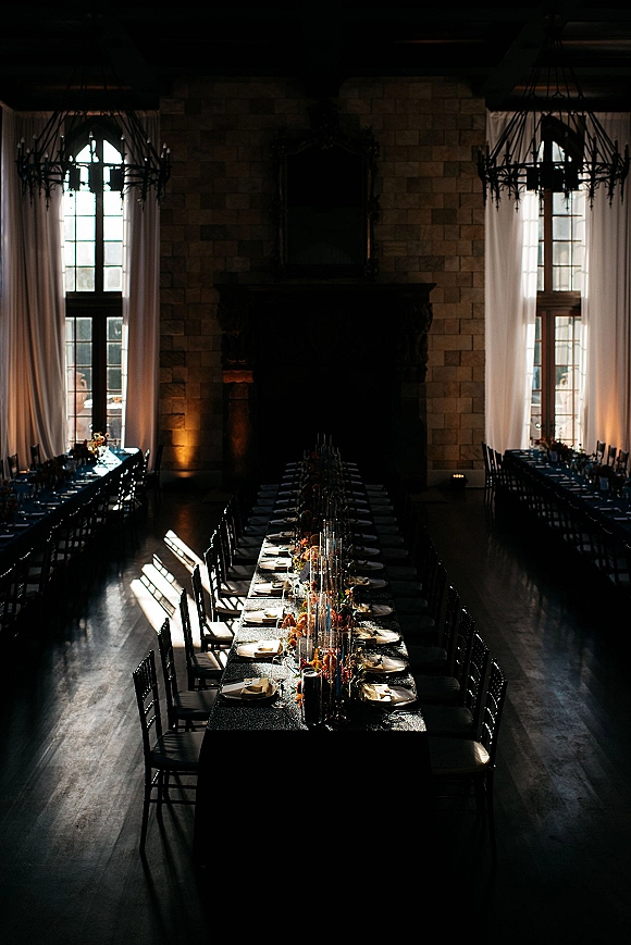 Reception tablescape with long banquet table wedding styling on black tablecloths, candles and garland centerpieces in a stone-walled hall