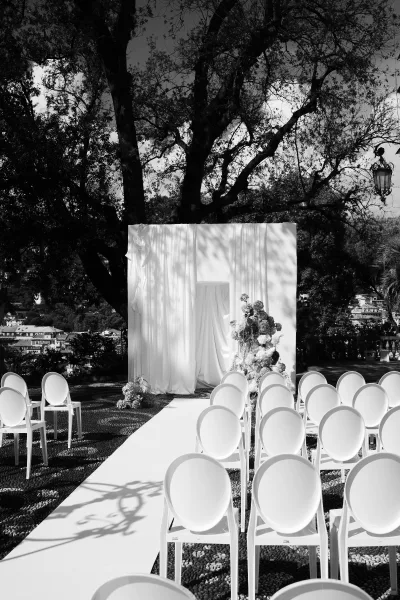 Ceremony setup for an outdoor wedding ceremony with a white aisle runner, white chairs, and draped fabric backdrop on a terrace under trees