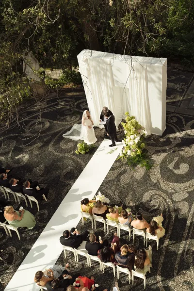 Ceremony setup with a white aisle runner leading to a white draped chuppah, viewed overhead in a mosaic-floor courtyard with greenery