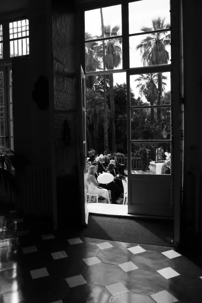 Ceremony moment at an outdoor wedding ceremony with guests seated under a white parasol, viewed from a doorway with palms and stone balustrade