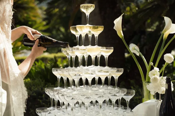 Champagne tower wedding coupe glass tower on a draped table with calla lilies and white flowers, set outdoors in sunlit palm greenery