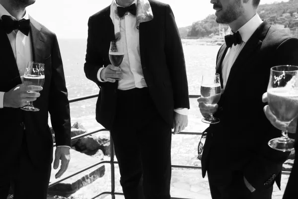 Groomsmen toast with champagne flutes in black tuxedos and bow ties on a balcony overlooking a rocky waterfront and hillside buildings