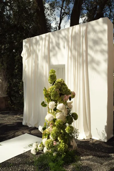 Wedding ceremony backdrop with draped wedding backdrop in white fabric, hydrangeas and greenery, set in a stone courtyard with trees and statue