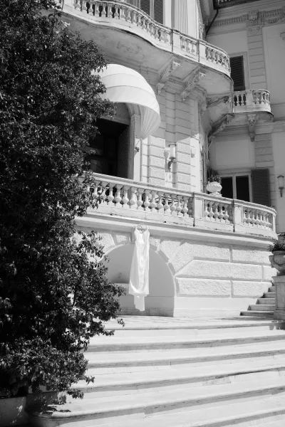 Wedding dress on hanger draped over a balcony railing beneath an awning, with a stone staircase and grand facade behind it