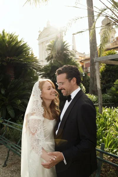 Couple portrait of bride and groom holding hands and laughing, lace mantilla veil and tuxedo in a palm-lined garden with sun flare