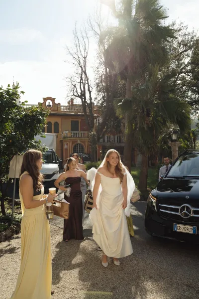 Bridal arrival as bride gets out of a car in an off the shoulder wedding dress with veil, bridesmaids nearby on a sunlit villa driveway with palm trees