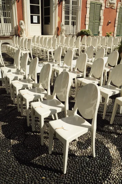 Ceremony seating with outdoor ceremony chairs in neat rows of white chairs with tags on a pebble courtyard beside a villa facade