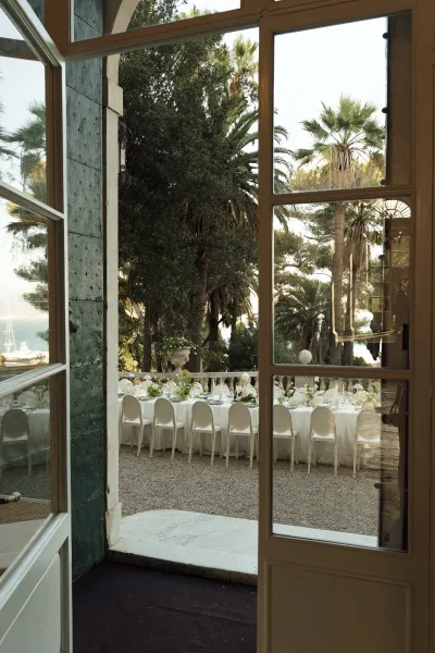 Reception tablescape with a long banquet table wedding setup, white linens and chairs, floral centerpieces, candles, and coastal terrace view