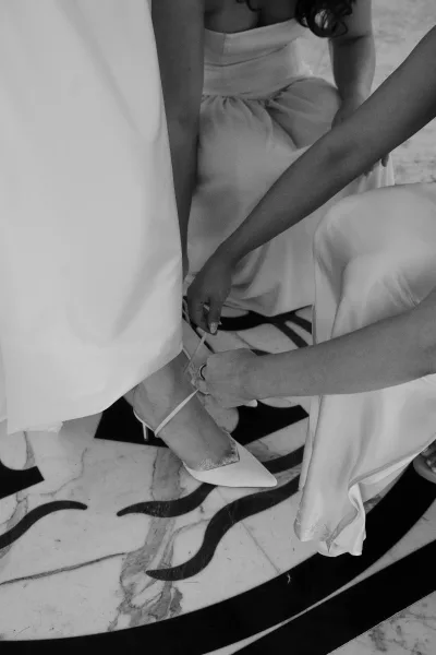 Bridal shoes as bride adjusts white ankle strap heels, bridesmaid nearby, ring visible on hand over geometric marble floor