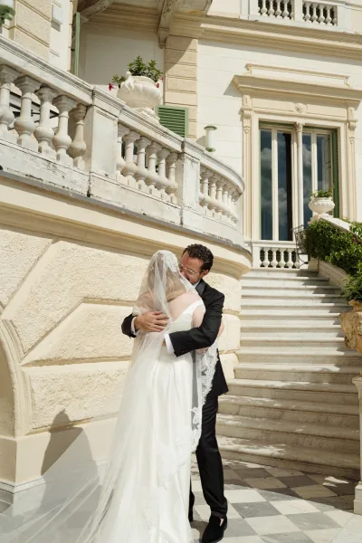 Wedding kiss as bride and groom embrace under a lace-trim veil on a stone staircase with villa balcony and shuttered windows