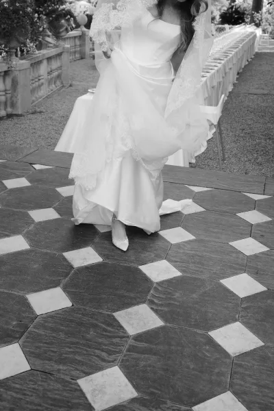 Wedding dress with off shoulder neckline and lace veil, bride holding hem beside pointed toe heels on a terrace by a long banquet table