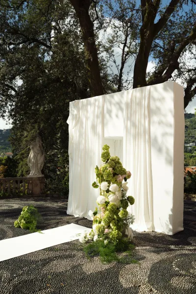 Wedding ceremony backdrop with white draping and greenery florals, set on a stone terrace with balustrade, statue, and hills beyond