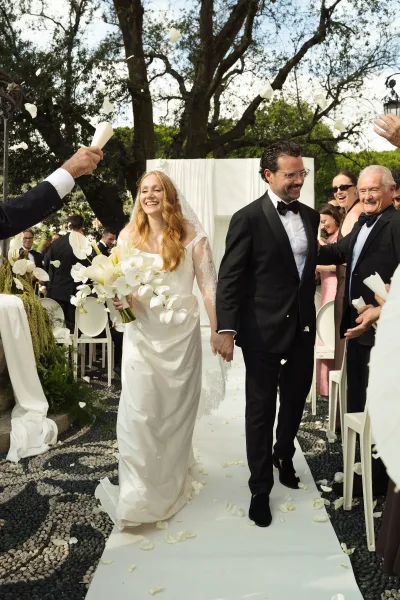 Wedding recessional as bride and groom walk aisle holding hands, with orchid bouquet and long veil, guests cheering in garden setting