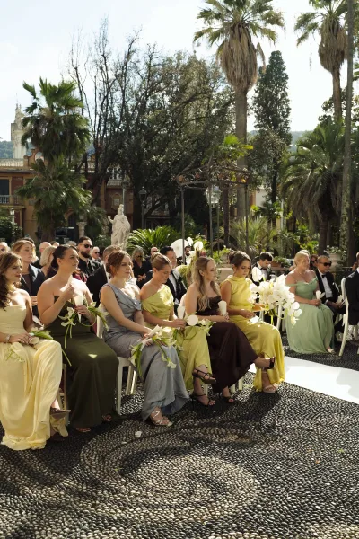 Ceremony guests in formal attire seated on white chairs, bridesmaids with calla lily bouquets in a sunny palm garden courtyard