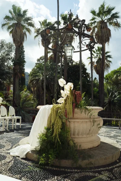 Ceremony backdrop with stone fountain, wrought iron canopy, and lantern above cascading anthurium florals in a palm-lined courtyard setting