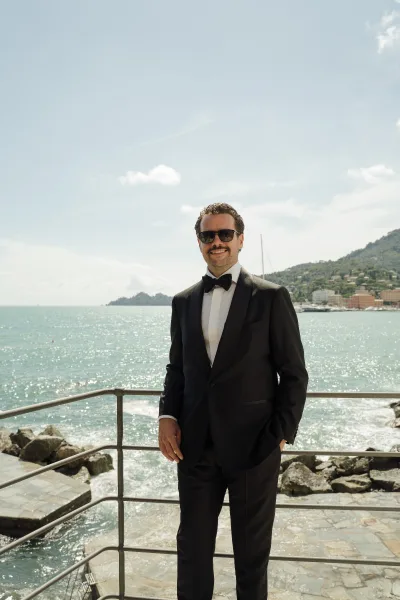 Groom portrait in a black tuxedo with bow tie and sunglasses, standing by an oceanfront railing above a rocky shoreline and sailboat