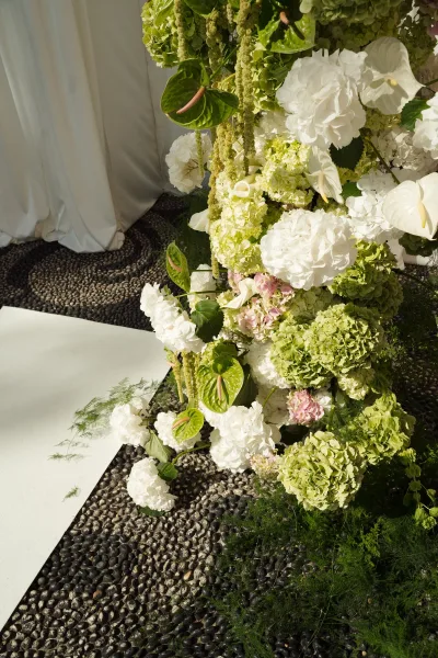 Wedding ceremony florals with ceremony aisle flowers, featuring hydrangeas, anthuriums, hanging amaranthus and greenery beside a white aisle runner on pebble floor