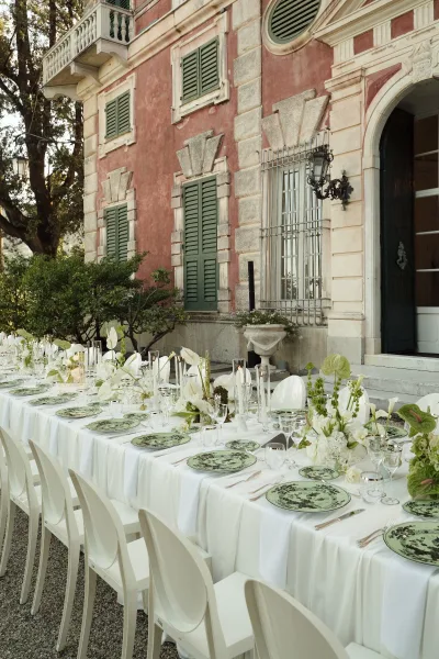 Reception tablescape with a long banquet table setup in white linen, green patterned plates, white florals and candlelight on a stone villa terrace