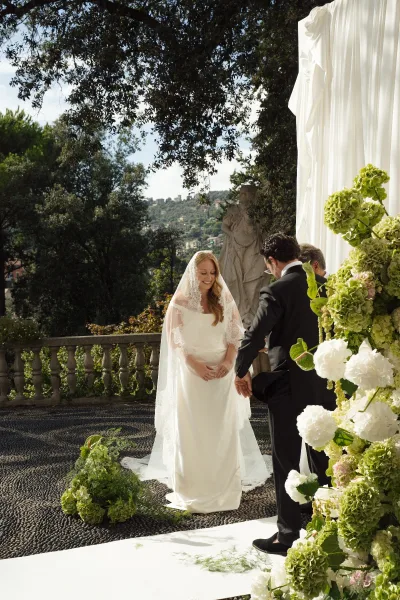Wedding ceremony moment as bride reads vows to groom beside officiant, framed by white draped backdrop and terrace hillside view